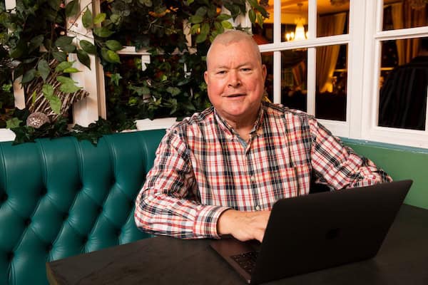 A friendly-looking man sits at a table with a laptop computer