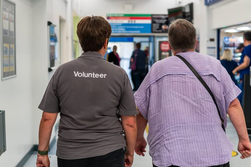 Two people viewed from behind walking down a hospital corridor. One wears a polo shirt with 'Volunteer' on it