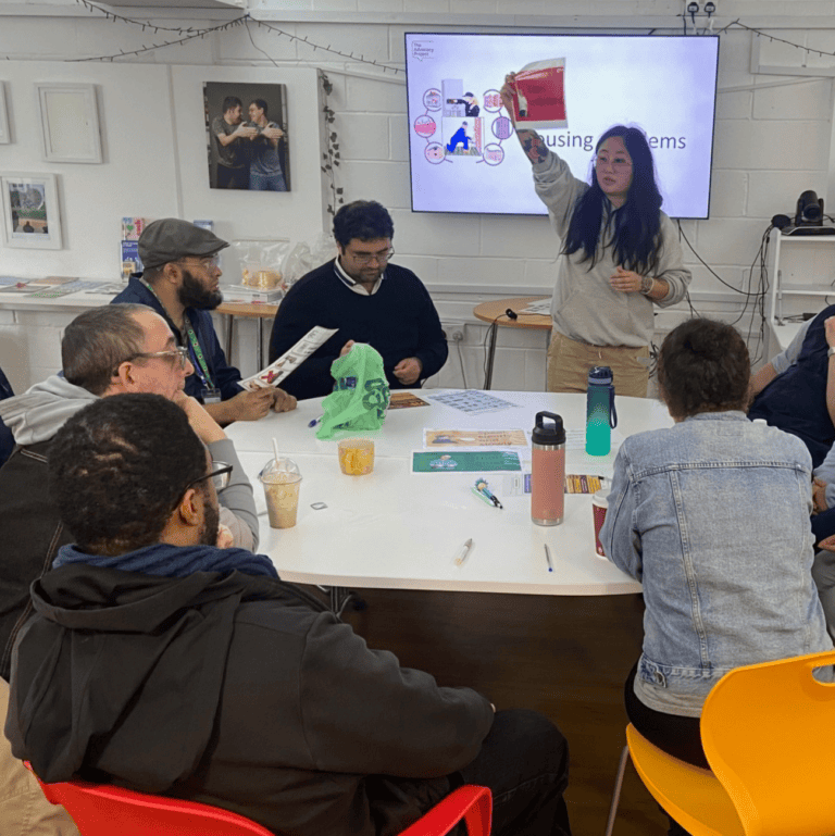 A group of LDN Community group attendees at a round table, sitting on brightly coloured chairs and facing a facilitator who is standing next to a large screen which is showing a presentation.