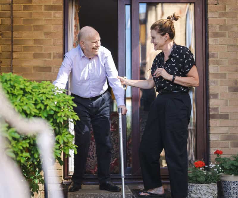 A older man with crutches walking out of a building smiles at a woman who is holding his arm.