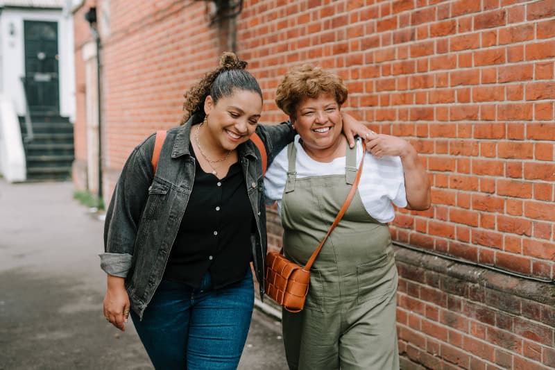 Two women walk down a street with their arms around each other smiling