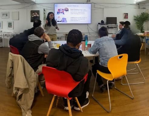A group of people sit at a table listening to a woman give a presentation