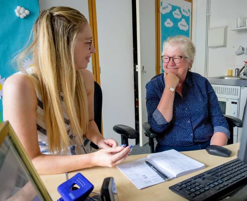 Two women - one younger, one older - sit at a desk in an office smiling at each other
